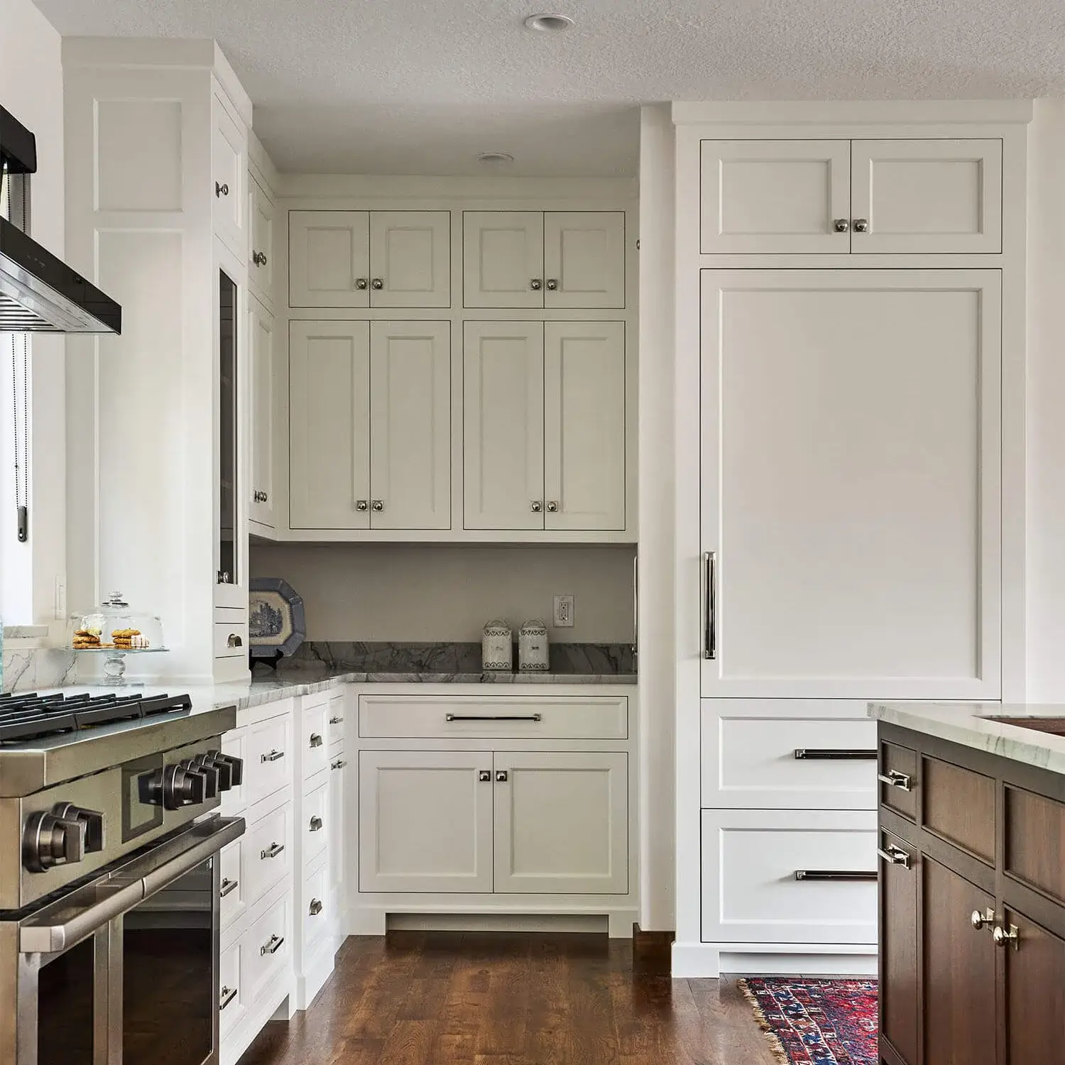 Luxury custom kitchen design featuring off white shaker kitchen cabinets with a flush inset face-frame structure. Includes a built-in integrated refrigerator panel, dark marble countertops, a professional stainless steel range stove, and polished chrome handles, contrasting with a dark stained wood island.