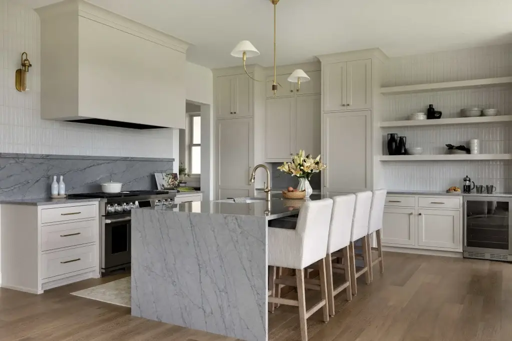 Side angle displaying modern greige kitchen cabinets behind a grey marble waterfall edge island with upholstered barstools.