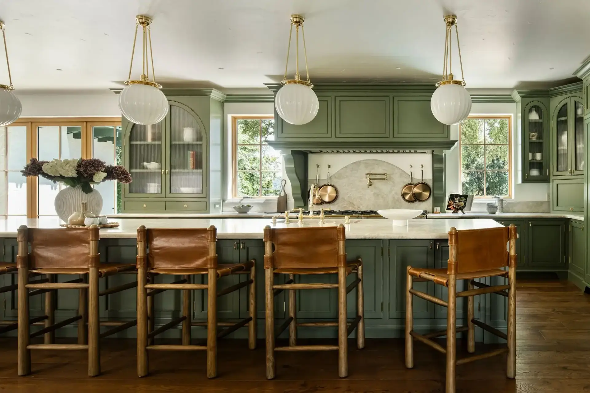 Wide view of dark green kitchen cabinets ideas featuring a large prep island with leather barstools, globe pendant lights, and arched display hutches.