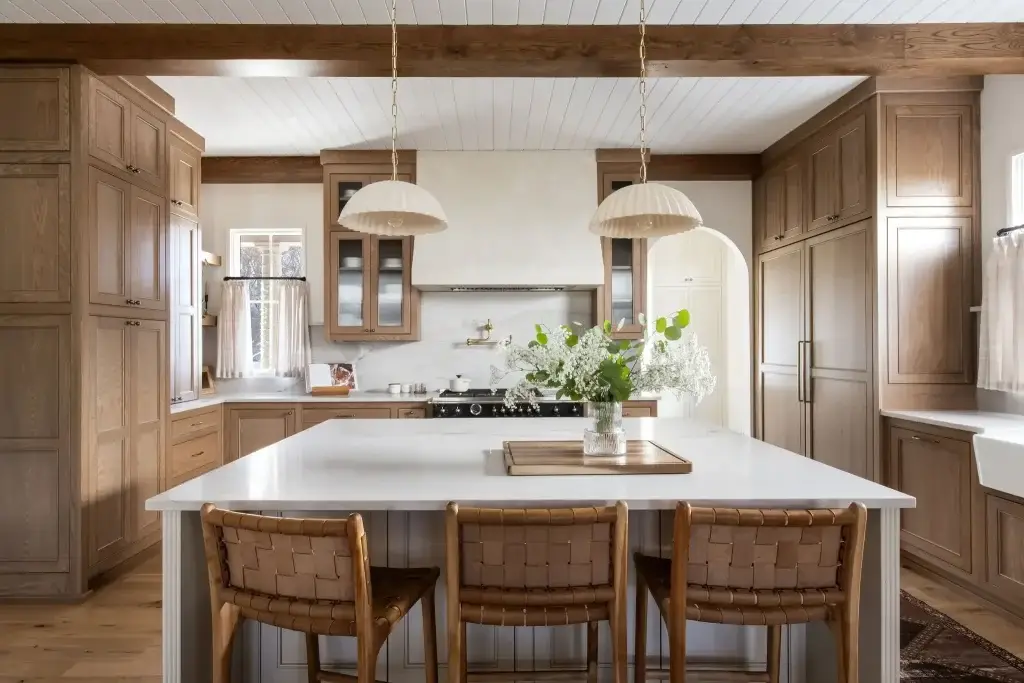 Symmetrical wide elevation of a honey oak modern kitchen with oak cabinets showcasing a white reeded island, leather stools, and plaster range hood.