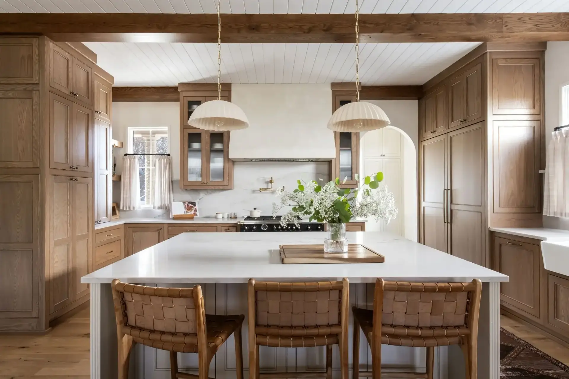 Symmetrical wide elevation of a honey oak modern kitchen with oak cabinets showcasing a white reeded island, leather stools, and plaster range hood.