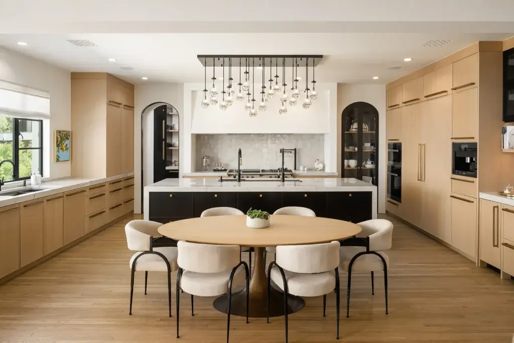 Symmetrical center view of rift cut white oak kitchen cabinets with black waterfall island, oval dining table, and raindrop chandelier.
