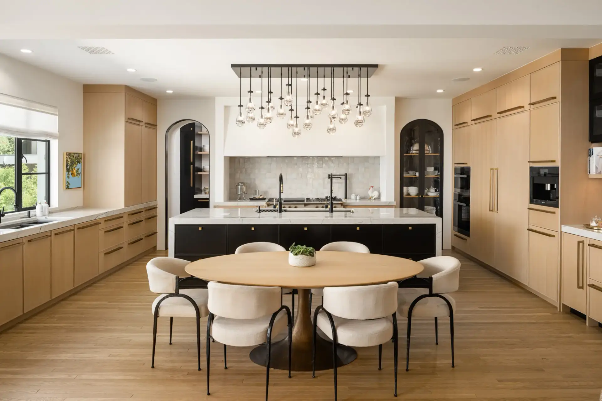 Symmetrical center view of rift cut white oak kitchen cabinets with black waterfall island, oval dining table, and raindrop chandelier.